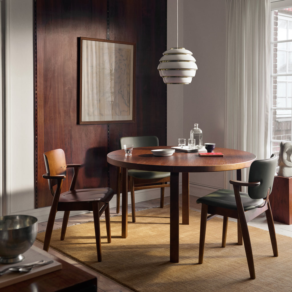 A dining room featuring a round, walnut‑stained 91 table, surrounded by Domus chairs. Above the table hangs an A331 pendant light, with framed artwork decorating the wall in the background.