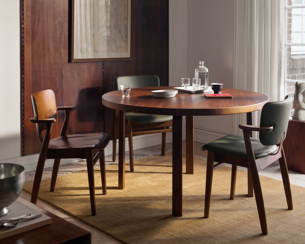 A beautiful dark‑wood wall forms the background of the room. In front of it stands a round, walnut‑stained 91 table surrounded by three Domus chairs.