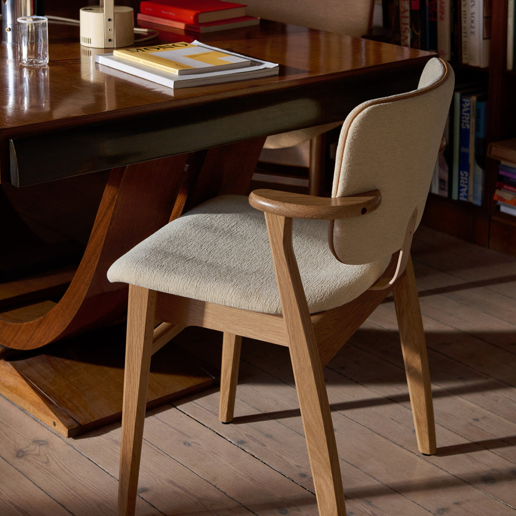 In a layered workspace with an old wooden desk, a Domus Chair with an oak frame is positioned beside the table, upholstered in Wow Re cream‑white fabric with Prestige Nougat leather piping.
