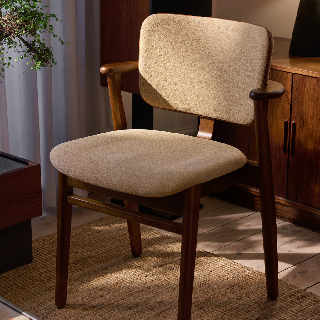 Domus Chair in walnut‑stained birch, upholstered in Wow Re beige‑pink fabric, shown in a layered living room with curtains and a sideboard in the background.