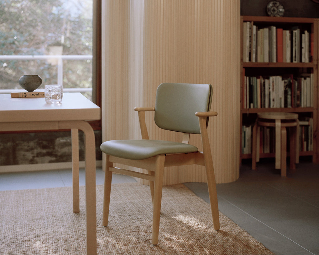 A minimalist room with a large window, where an Aalto Screen 100A stands beside a rectangular Aalto table. Next to the table, a honey‑stained Domus Chair upholstered in Spectrum Leaf leather with Spectrum Moss piping is shown from the side, while a library room appears softly in the background.