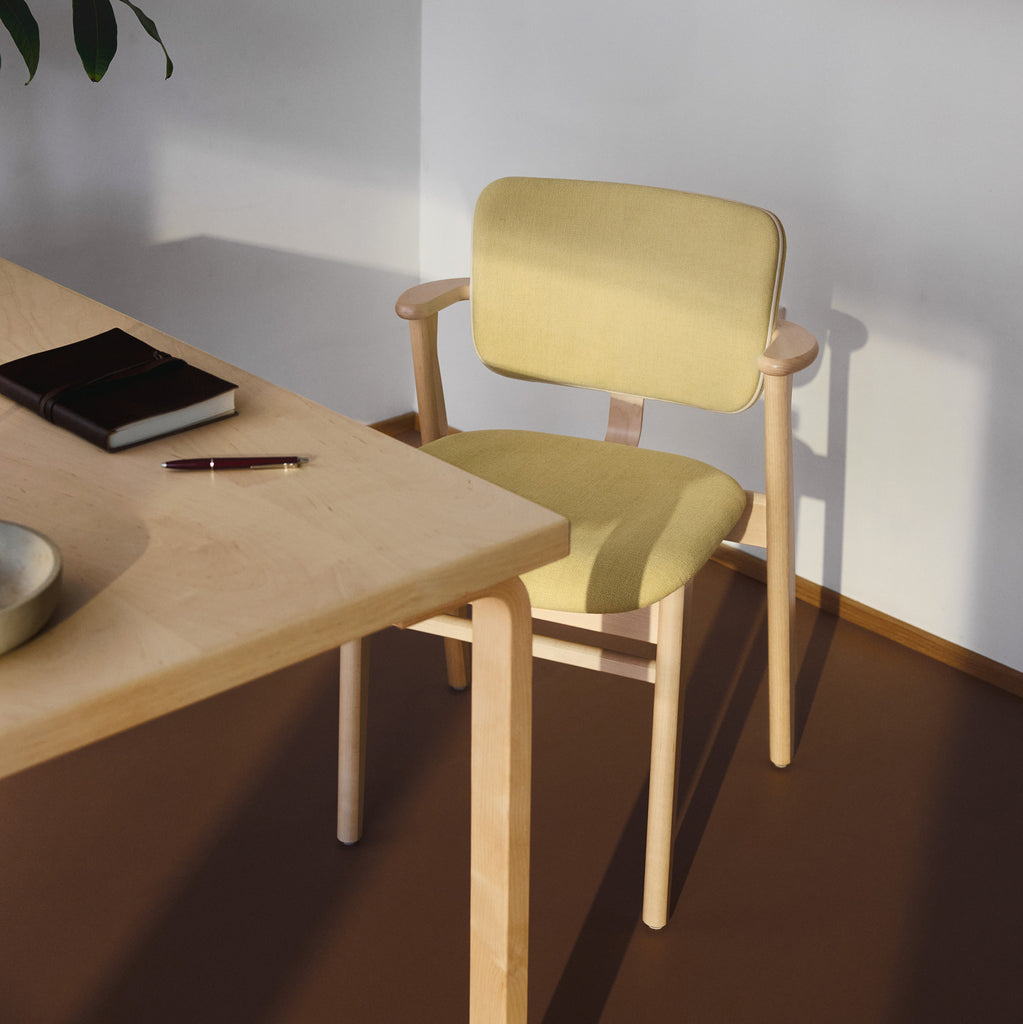 A Nordic‑style room with a clear‑lacquered birch Domus Chair upholstered in Hola Canary fabric and finished with Prestige Cream leather piping, placed beside a table.