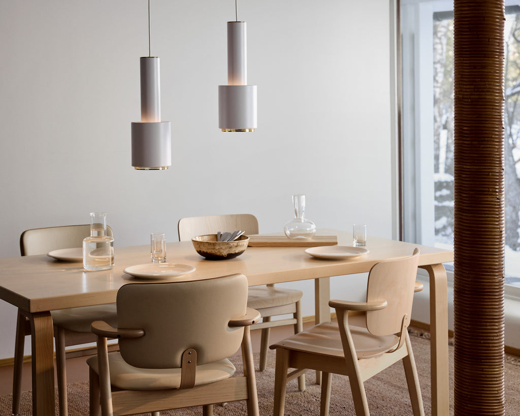 Nordic‑style dining room featuring Alvar Aalto’s rectangular dining table surrounded by four Domus chairs, with two white A110 pendant lights with brass rings hanging above the table.