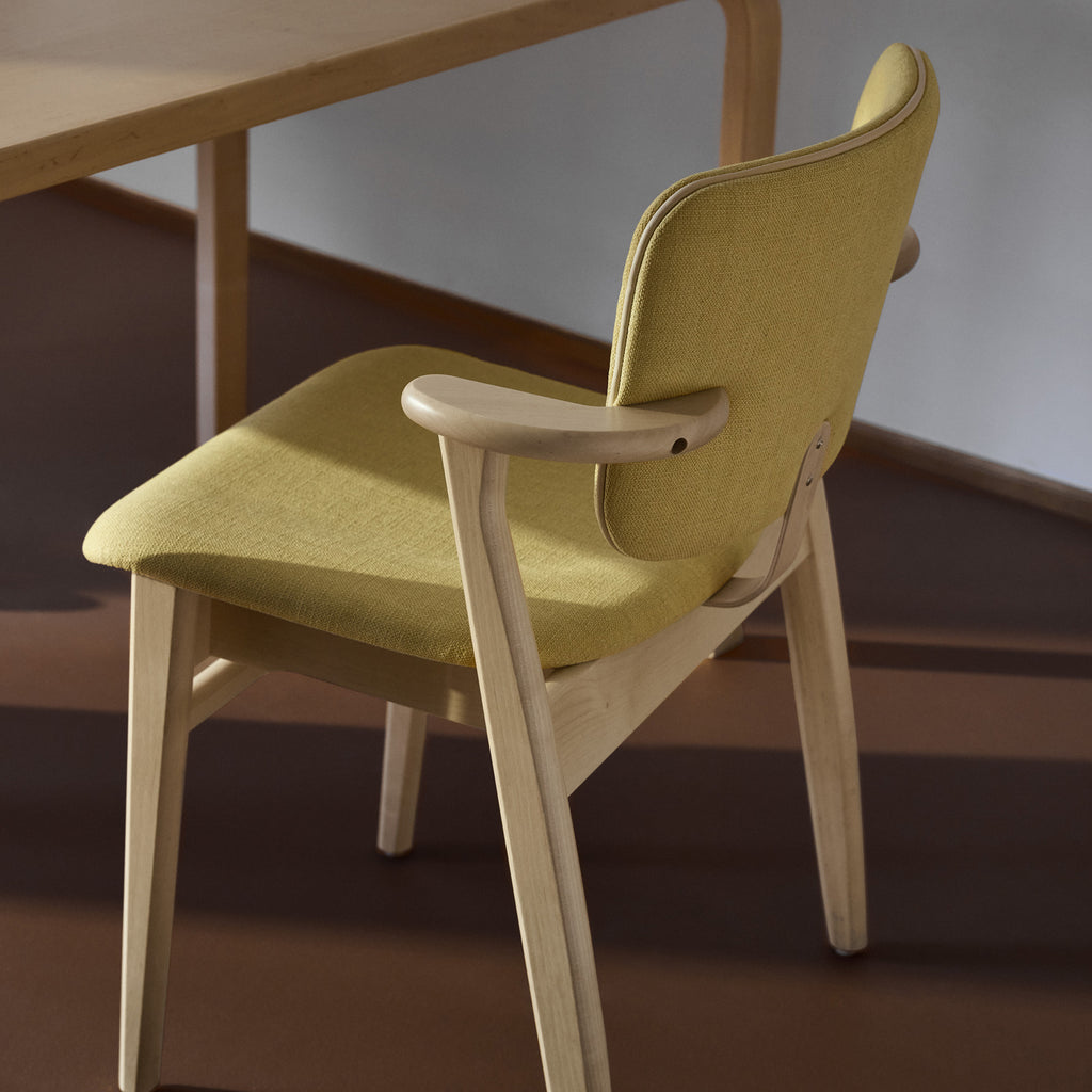 Side‑rear angled view of the Domus Chair, highlighting its elegant curved birch frame, the light‑yellow Hola Canary upholstered backrest and seat, and the refined Prestige Cream leather piping.