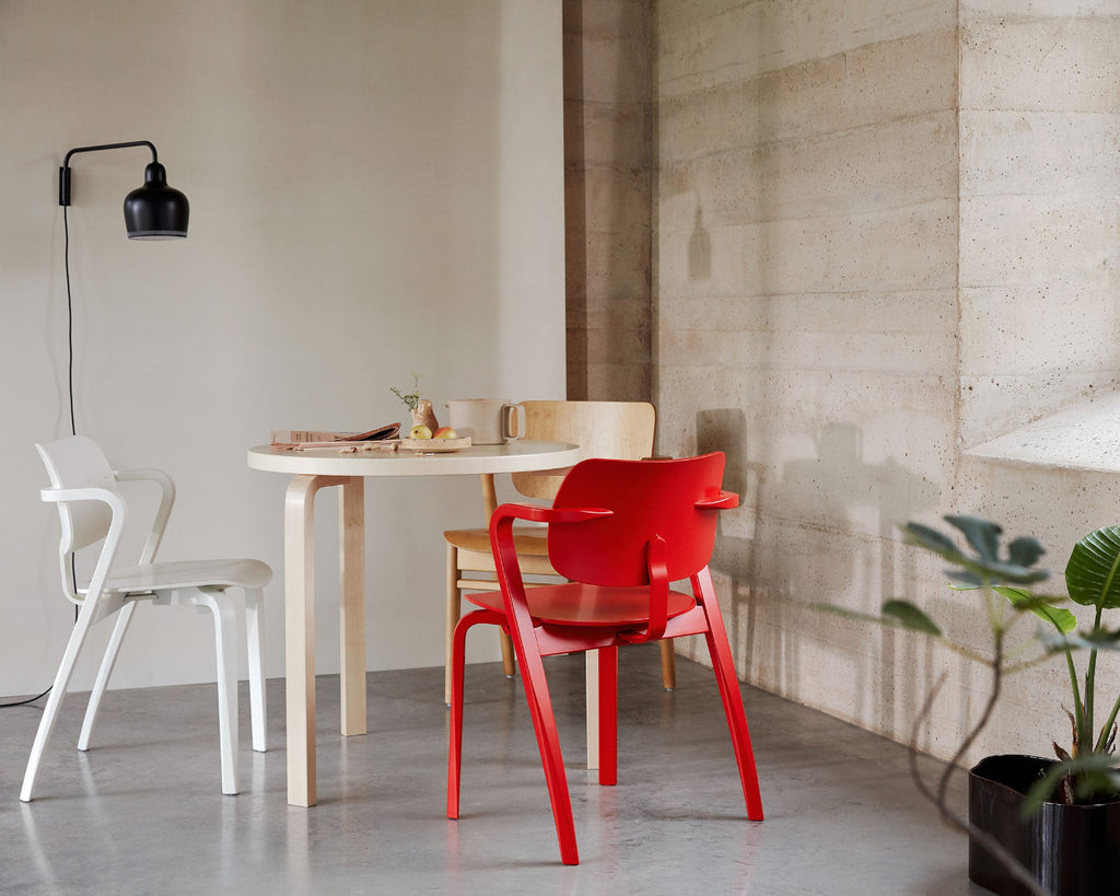 In a highly minimalist space, round birch Artek Table 90B with two Aslak Chairs, one white and one red. Part of a honey-stained Domus Chair is visible behind the table, and a black A330S Wall Light is mounted on the wall.