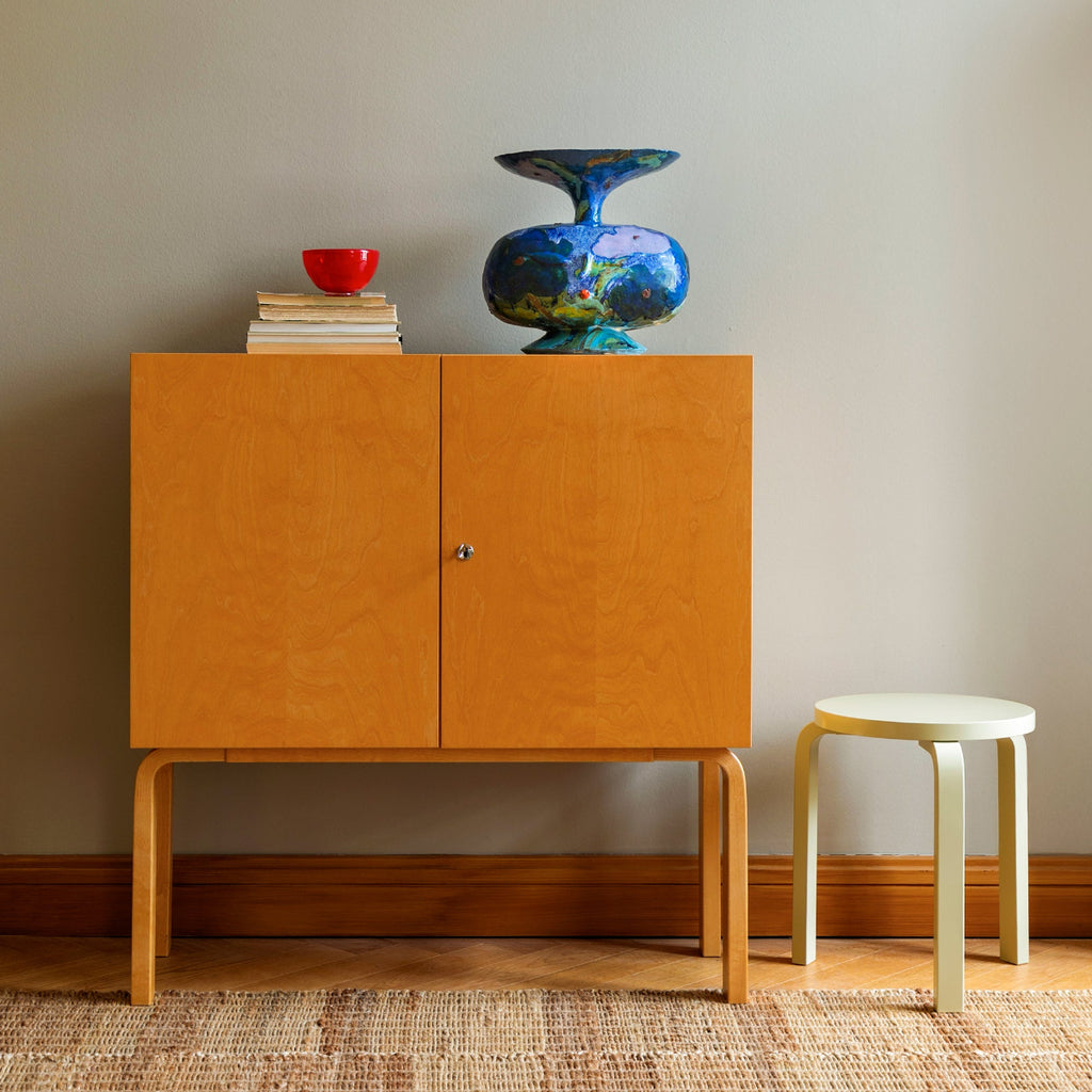 Interior photograph featuring a Cabinet 250 placed against a wall with a Stool 60 positioned beside it. An artwork and a stack of books are displayed on top of the cabinet.