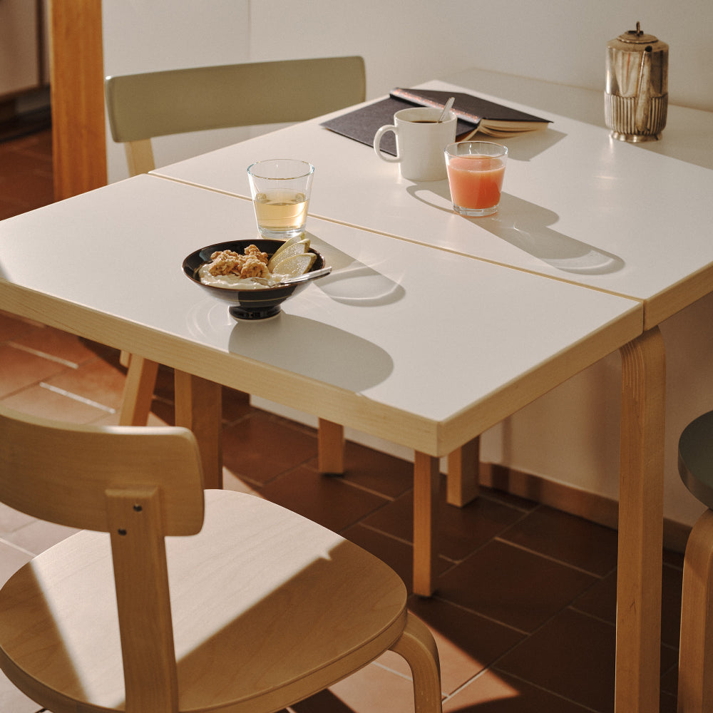 Close-up interior view of a small dining area next to a kitchen, featuring Aalto Foldable Table DL81C with a white laminate top. Two Chair 69 and one Stool 60 are placed around the table, creating a compact and functional dining setup.