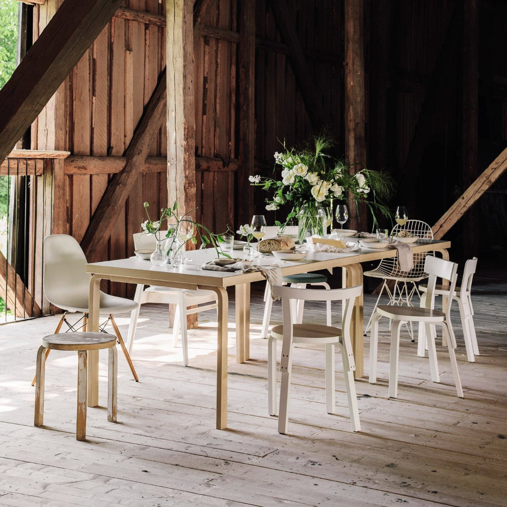 Interior view of a dining group located in a barn, featuring rectangular Aalto Table 82A surrounded by different white lacquered seating, including Stool 60 and Chairs 65, 66, 68, and 69.