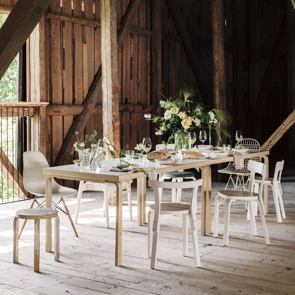 Interior view of a dining group located in a barn, featuring a rectangular Aalto Table 82A surrounded by white lacquered seating, including Stool 60 and Chairs 65, 66, 68, and 69.