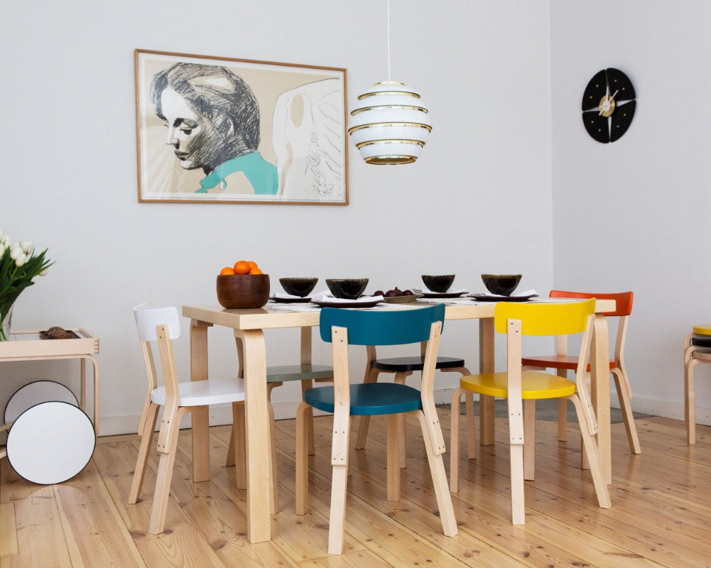 Interior view of a dining area featuring rectangular Aalto Table 82B surrounded by differently coloured Chair 69. A Tea Trolley 900 and a stack of Stool 60 are placed against the wall, while Pendant Light A331 “Beehive” hangs above the table.