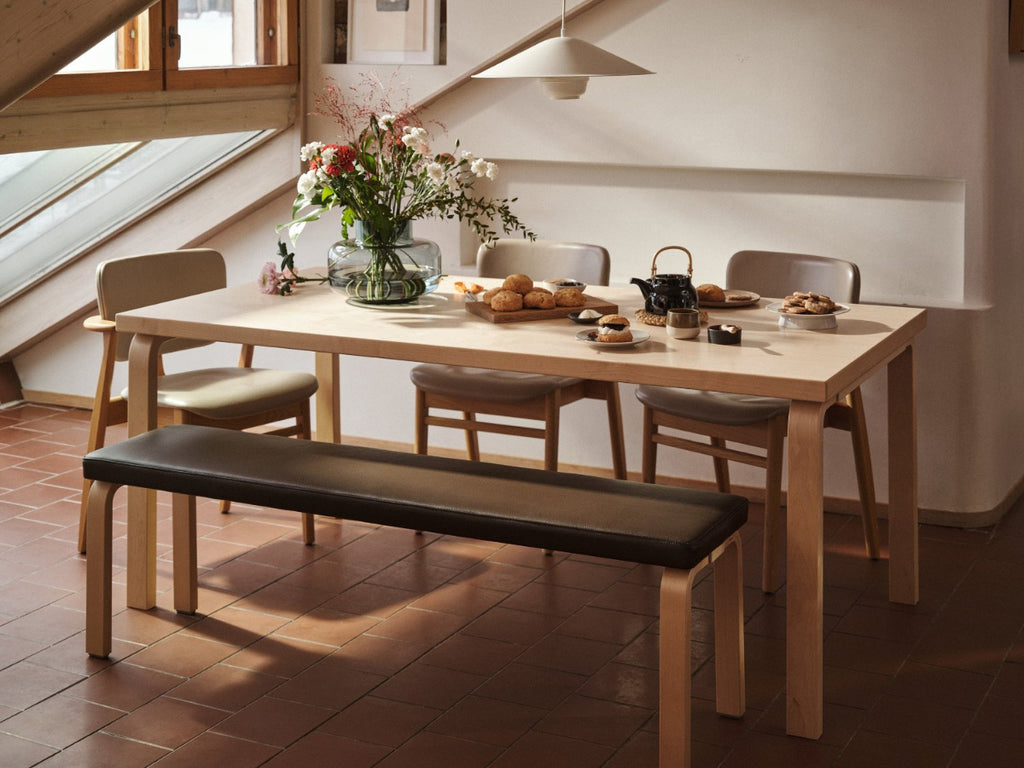 Interior view of a dining area featuring Aalto Table 83 with a leather‑upholstered Bench 168B along one side. Three Domus Chairs are placed around the table. Kori Pendant Light with Dune Shade hangs above, and the table is set with a large bouquet of flowers and food.