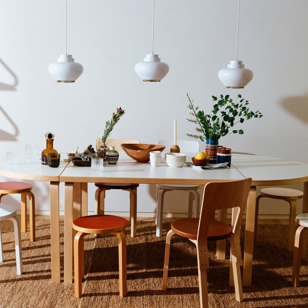 A dining space featuring an oval table arrangement formed from Aalto tables. Around the table are 60 Stools and 66 Chairs. Above the table hang three Pendant Lights A331 “Turnip”. The table is set with decorative objects and tableware.