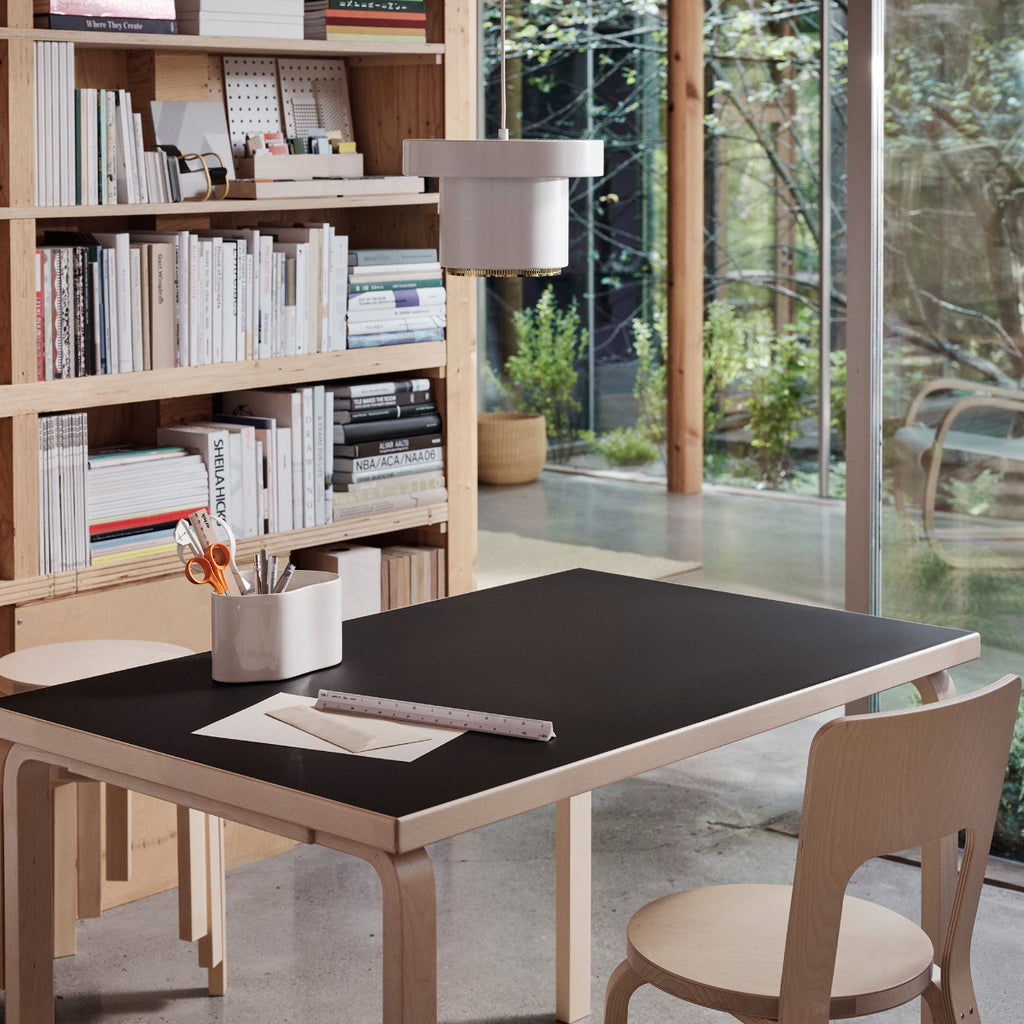 Home office interior featuring a rectangular Aalto Table with a black linoleum top placed in the centre of the room, with Chair 66. A Riihitie Plant Pot is on the table, and Pendant Light A201 hangs above. A large bookcase is visible in the background.