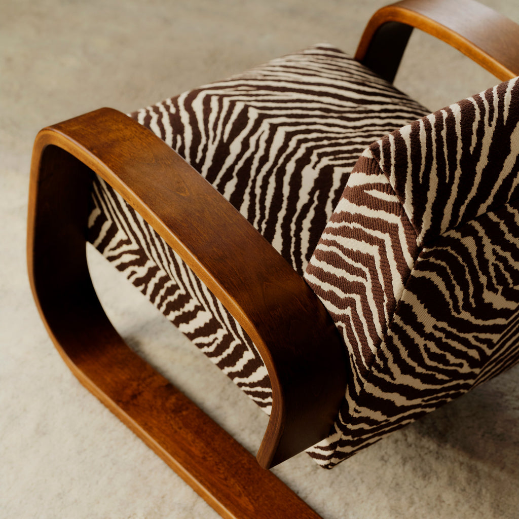 Top-down close-up photograph of an Artek Armchair 400 upholstered in brown and white Zebra fabric. The armchair features walnut-stained armrests.