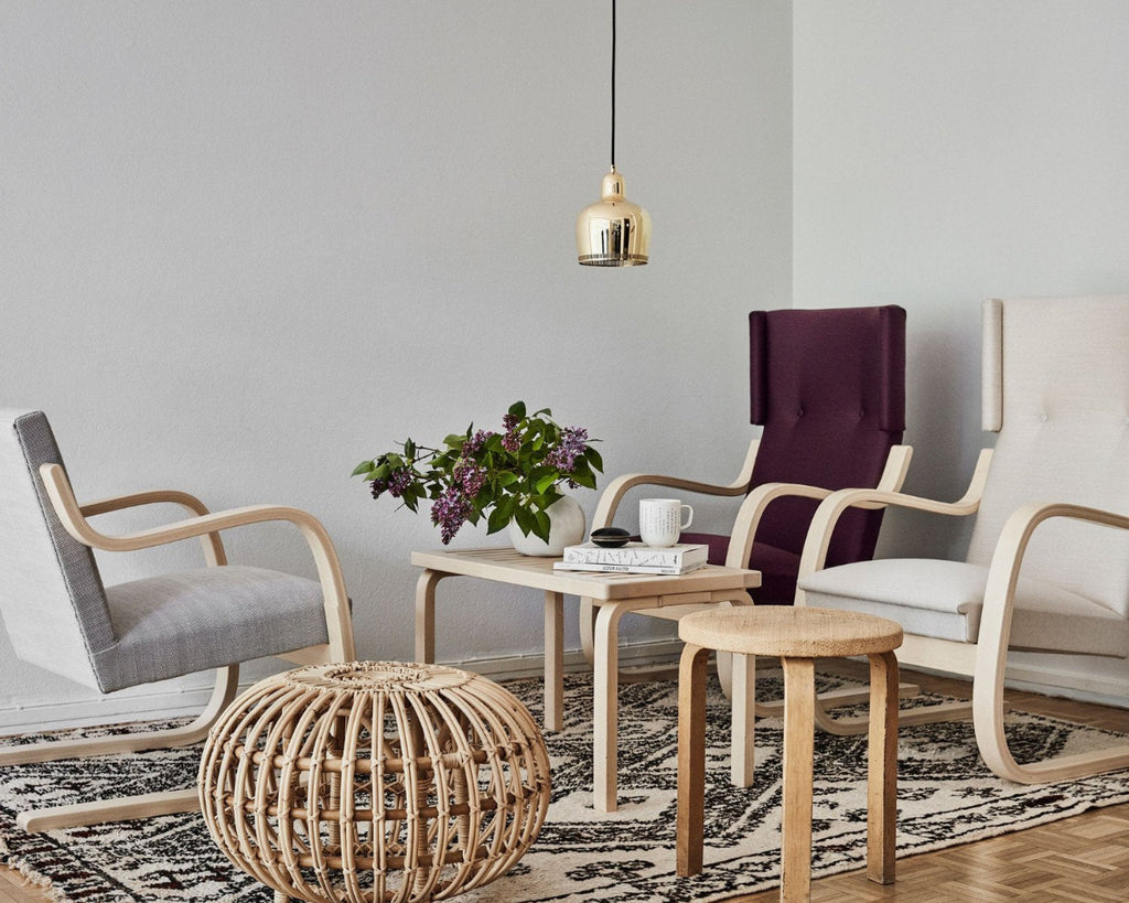 Living room with two Armchair 401 placed side by side, one in lilac upholstery and one in a light tone. On one side is Armchair 402 upholstered in grey fabric. Between the armchairs is birch Bench 153B with slatted top, styled with flowers and books, accompanied by a patinated Artek 2nd Cycle Stool 60. The seating group rests on a Moroccan rug, and above the bench hangs Pendant Light A330S “Savoy”.