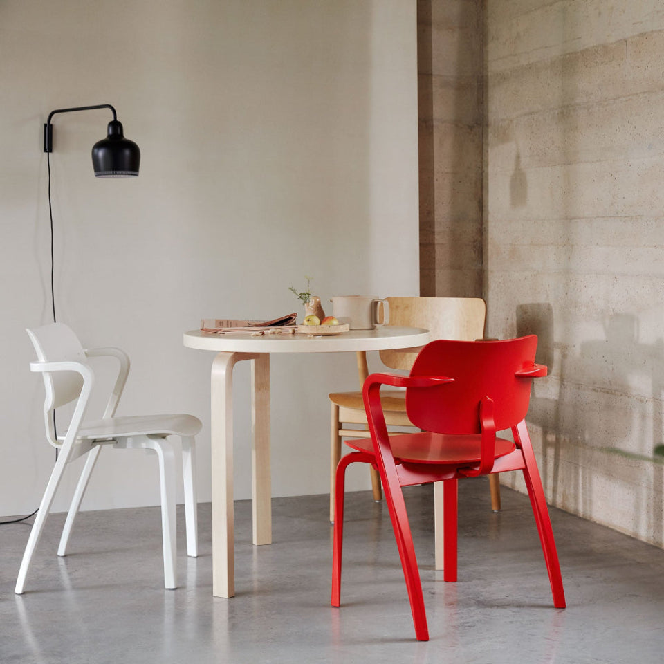 In a highly minimalist space, round birch Artek Table 90B with two Aslak Chairs, one white and one red. Part of a honey-stained Domus Chair is visible behind the table, and a black A330S Wall Light is mounted on the wall.