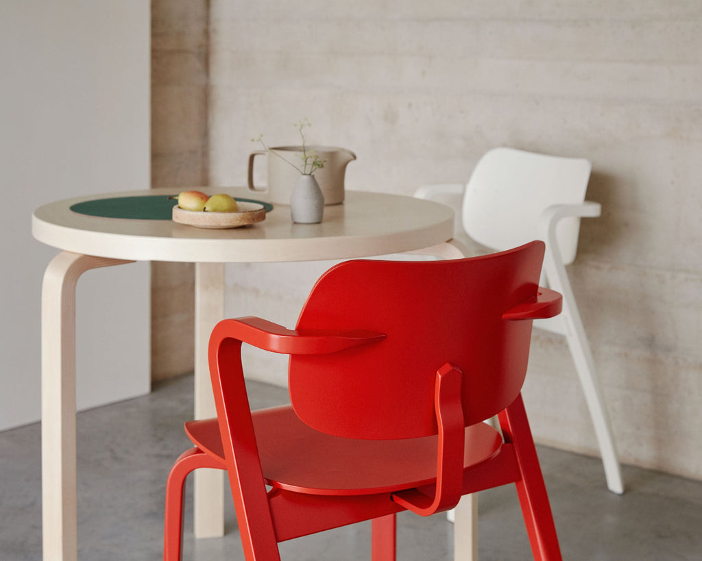 In a highly minimalist space, close-up view from behind of red Aslak Chair. Birch Artek Table 90B and white Aslak Chair visible in background.