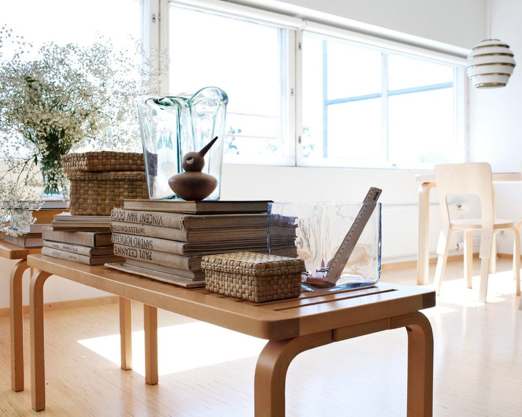 Two birch Bench 153A with slatted tops are placed lengthwise, styled with books, small objects, and a vase with white flowers. Large windows are visible in the background along with an Artek Table and Chair 66. Above the table hangs Pendant Light A331 “Beehive”.