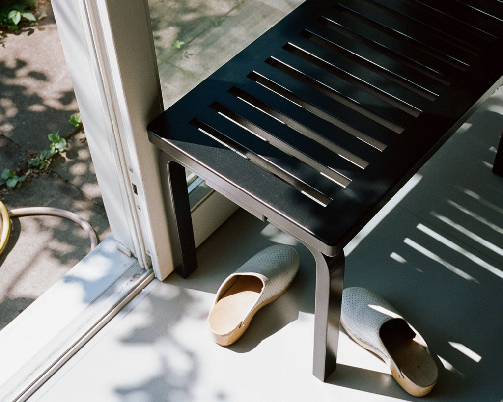 Top-down close-up view of black Bench 153B with slatted top, partially visible. Two wooden shoes are placed underneath the bench. The bench is positioned in front of a large window next to an exterior door, with tiled surfaces visible through the windows.