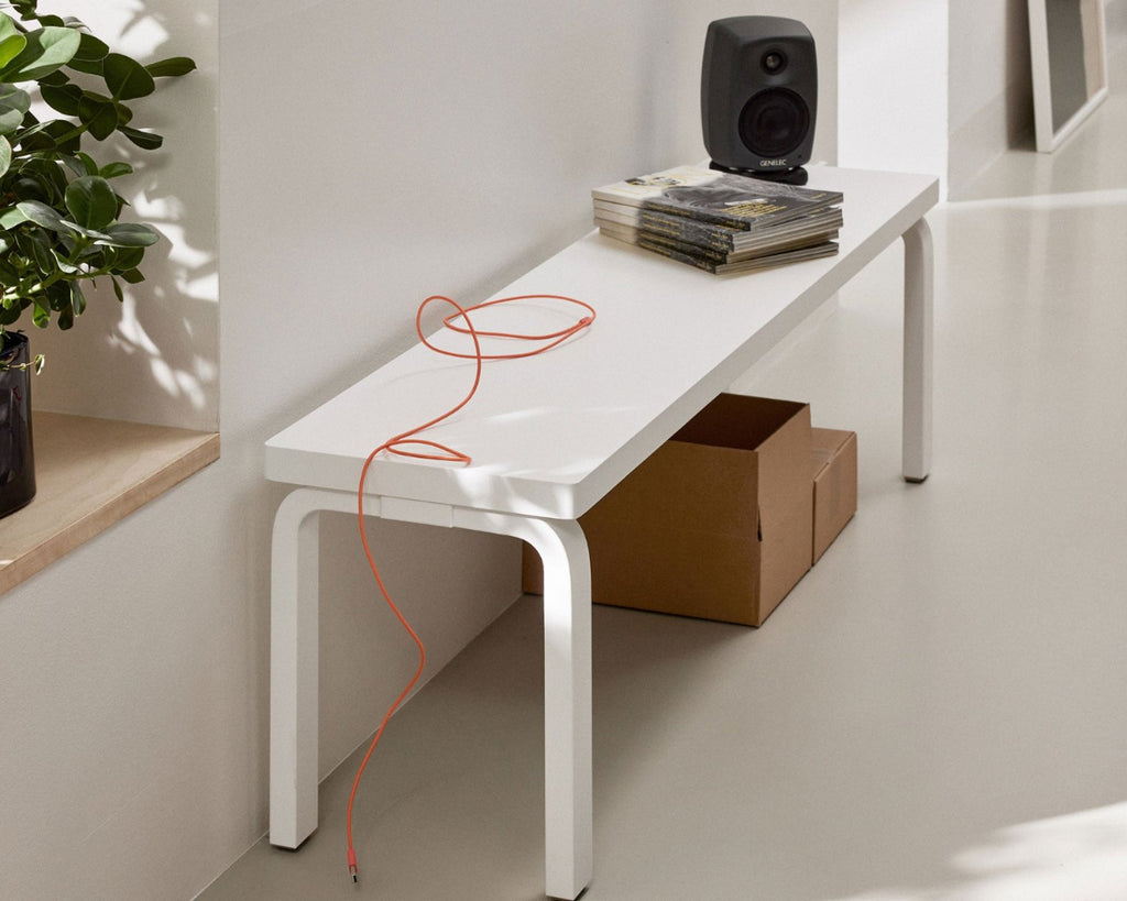 In a light-colored hallway, a white Bench 168B is placed against the wall. On top of the bench are an orange cable, a stack of magazines, and a stereo, with cardboard boxes stored underneath.