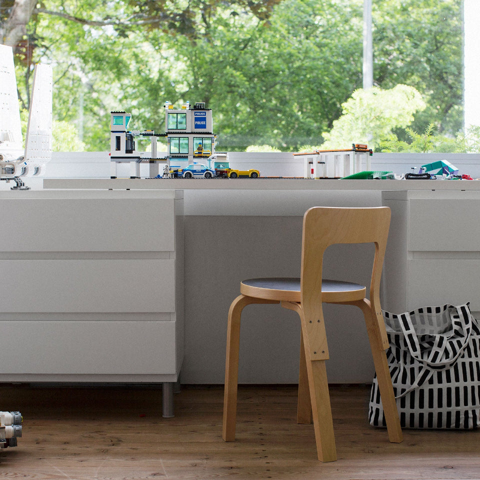 Children’s room with a white desk featuring drawers on both sides placed under a large window. Under the desk is Artek Chair 65 with a black linoleum seat. Next to the chair is a black-and-white Siena fabric bag, and Lego constructions are arranged on the desktop.