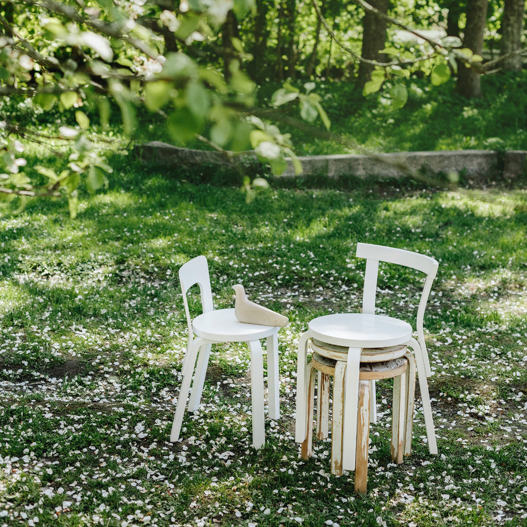 Outdoor interior view on a lawn featuring a white lacquered Chair 65. Next to it, two white Artek 2nd Cycle Stool 60 are stacked, with a white lacquered Chair 68 placed on top of the stools.