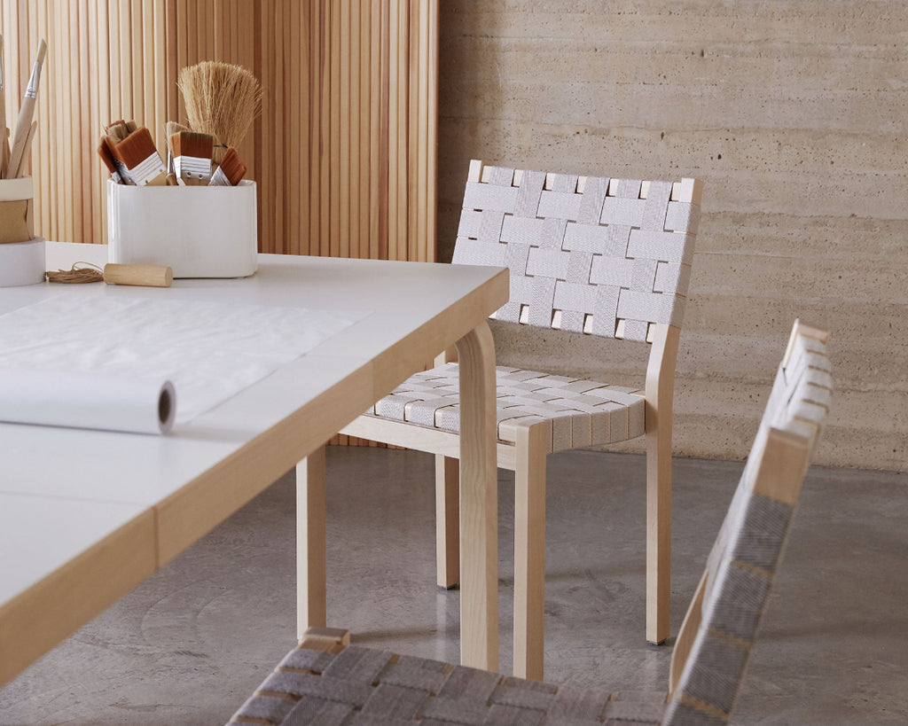 Minimalist workspace featuring Artek Extension Table 97 with a white laminate surface, paired with two Chair 611. In the background, Screen 100B is partially visible. On the table, Riihitie Plant Pot Medium B in white holds paintbrushes, and a large white paper roll is partially unfolded across the tabletop.
