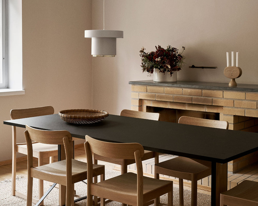 Dining area with rectangular Artek Kaari Table surrounded by six oak Atelier Chairs. White A201 Pendant Light hangs above the table. Behind the table is a brick fireplace topped with a white Riihitie Plant Pot with flowers and Lucia Candleholder.