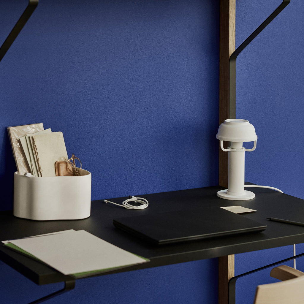 Interior view featuring white Kori Table Light placed on Kaari Wall Shelves mounted on a dark blue wall. A white Riihitie Plant Pot small is also on the shelf.