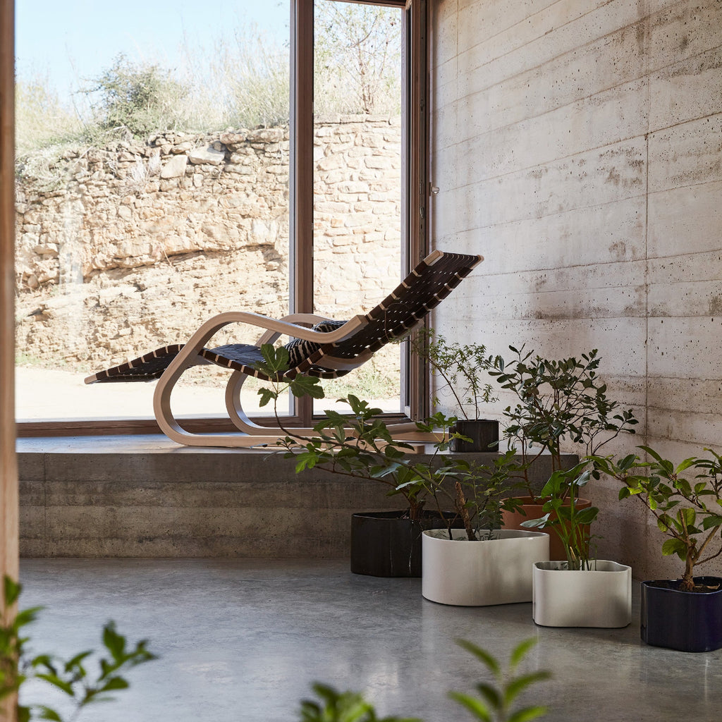 Minimalist living room featuring Lounge Chair 43 placed on a raised platform in front of a large window. Beneath the platform, Riihitie Plant Pots in different sizes contain plants.