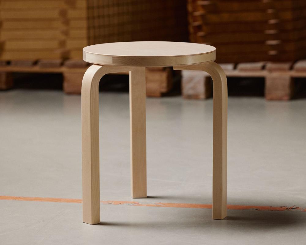 Making‑of view featuring Artek Stool 60 with a clear lacquer finish, standing on a factory floor. In the background, stacks of Artek products are visible in storage boxes.