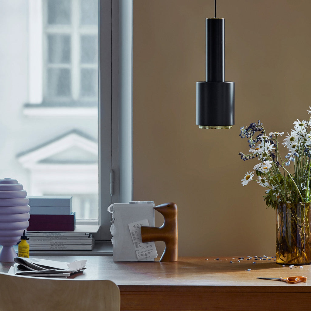 Interior view of a workspace lit by daylight, featuring a black Pendant Light A110 “Hand Grenade” with a brass ring above a desk. The workspace is positioned under a window, with an old apartment building visible outside.