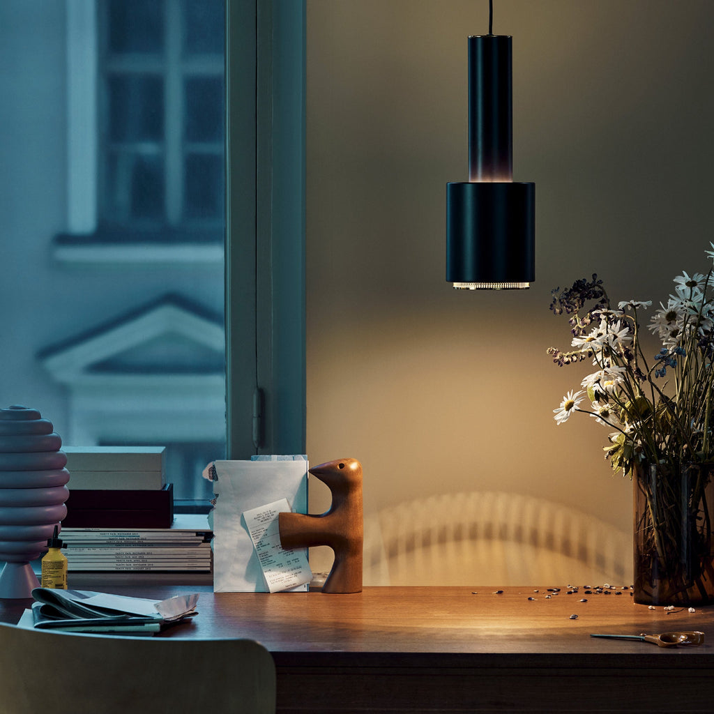 Interior view of a dimly lit workspace featuring a black Pendant Light A110 “Hand Grenade” with a brass ring above a desk. The workspace is positioned under a window, with an old apartment building visible outside.