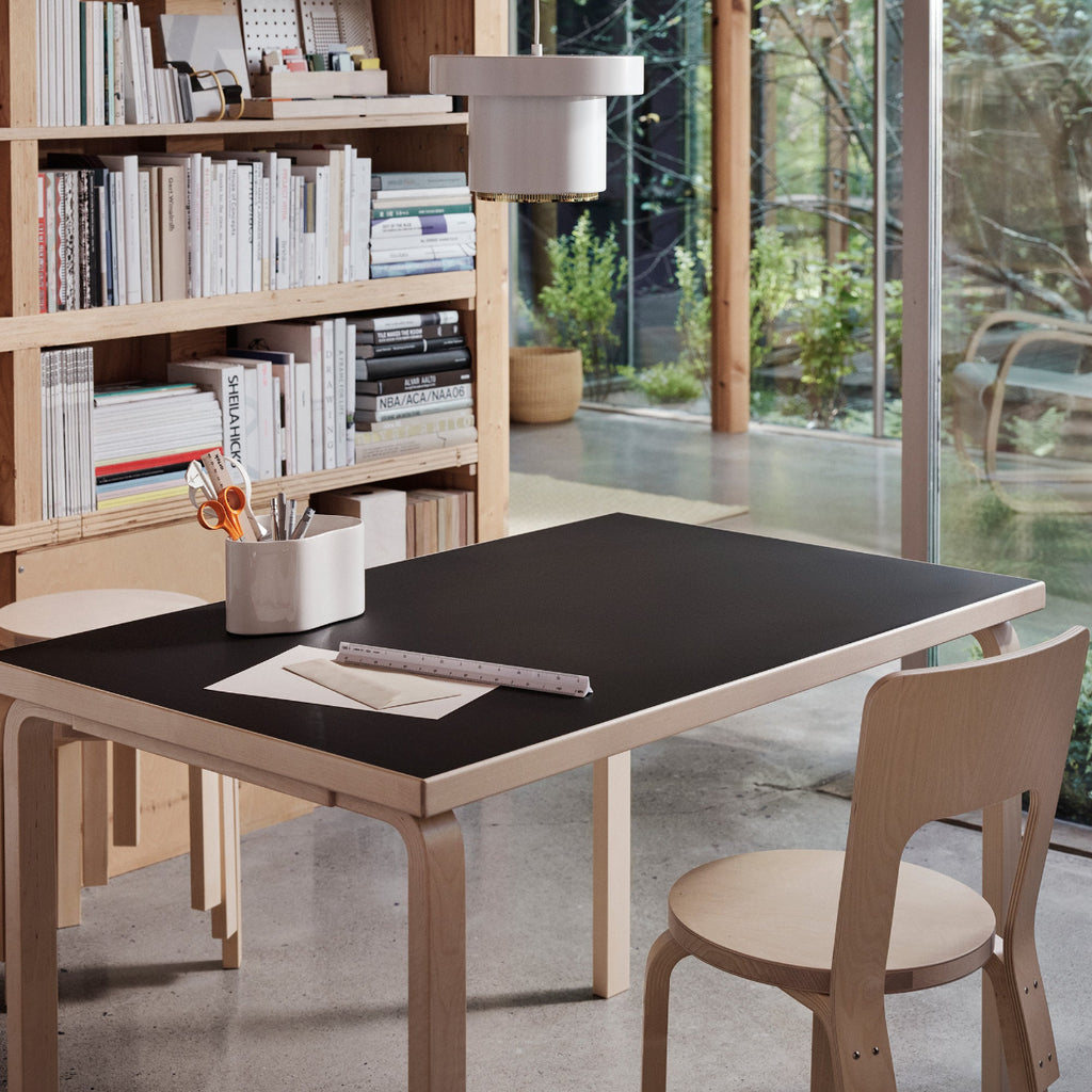 Home office interior featuring rectangular Artek Aalto Table 82B with a black linoleum top placed at the center of the room. Artek Chair 66 in birch is used as a workspace chair, with a large bookcase visible in the background. Pendant Light A201 hangs above the table.
