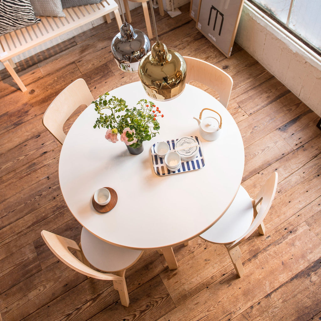 Top-down view of a dining area featuring a round Artek Aalto Table 90A surrounded by four Chair 66. Two Pendant Lights A330S “Golden Bell” hang above the table, and a slatted Bench 153A with cushions is visible in the background.