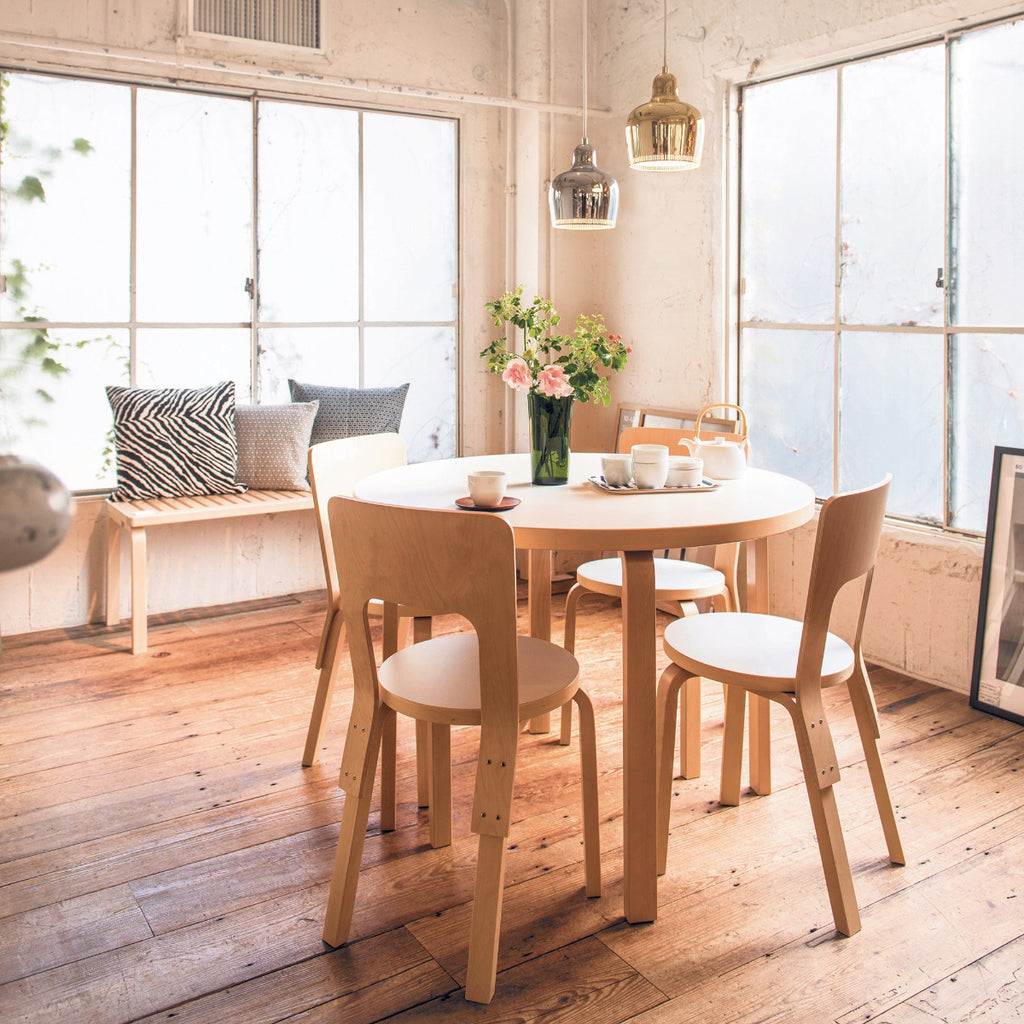 Dining area featuring a round Artek Aalto Table 90A surrounded by four Chair 66. Two Pendant Light A330S “Golden Bell” hang above the table, while a slatted Bench 153A with cushions is visible in the background.
