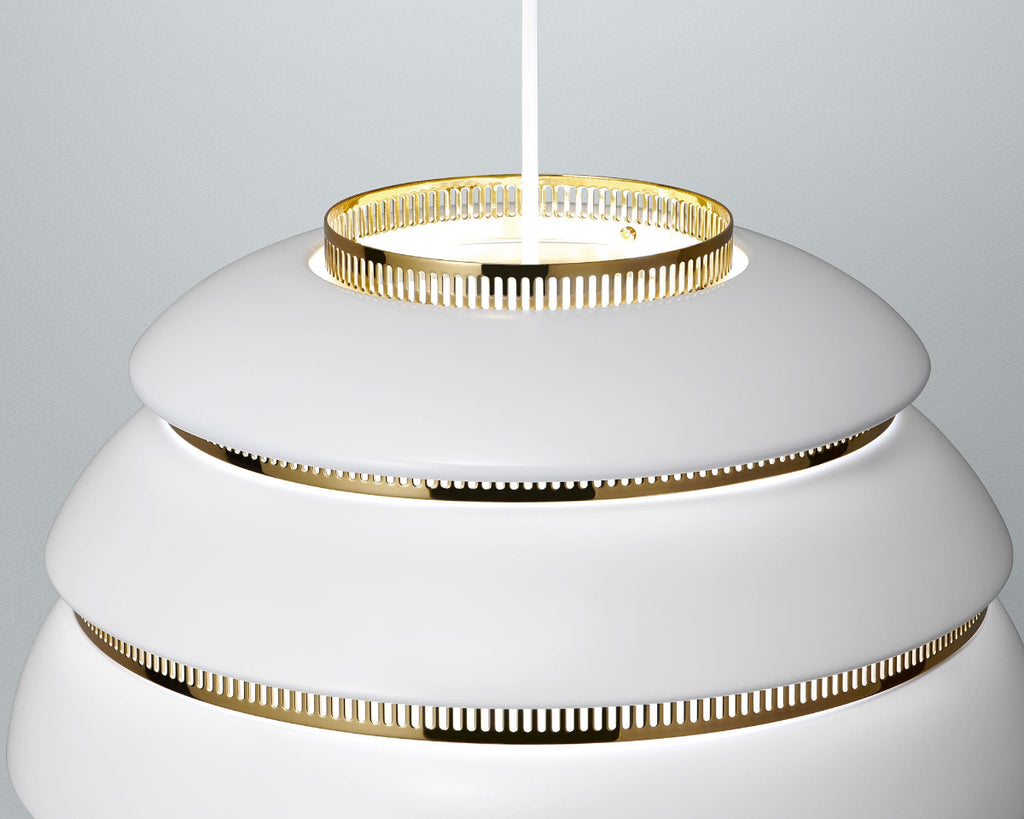 Close-up product image of the upper part of white Artek Pendant Light A331 “Beehive” with brass details. The pendant light is switched on and photographed against a grey background.