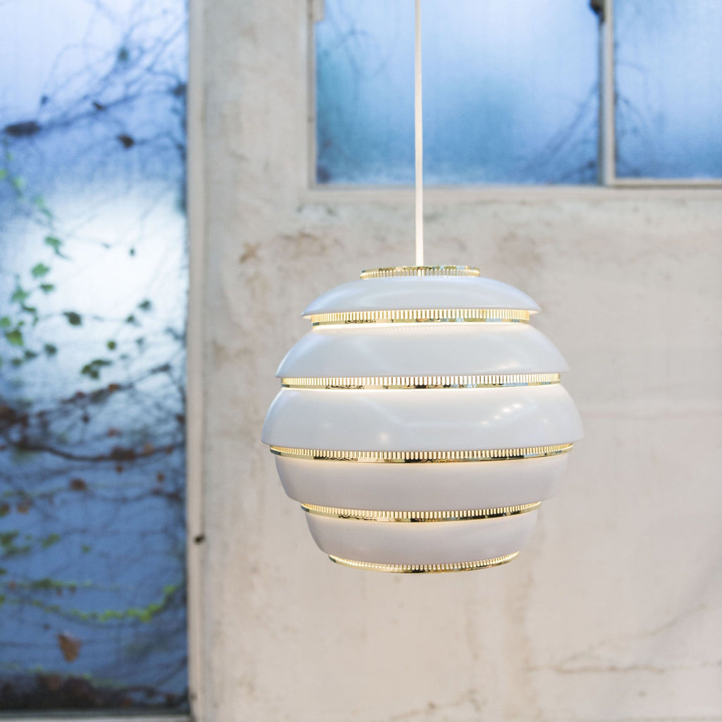 Minimalist interior featuring a white and brass Pendant Light A331 “Beehive” hanging in the space. Large windows reveal an outdoor view during the blue hour.