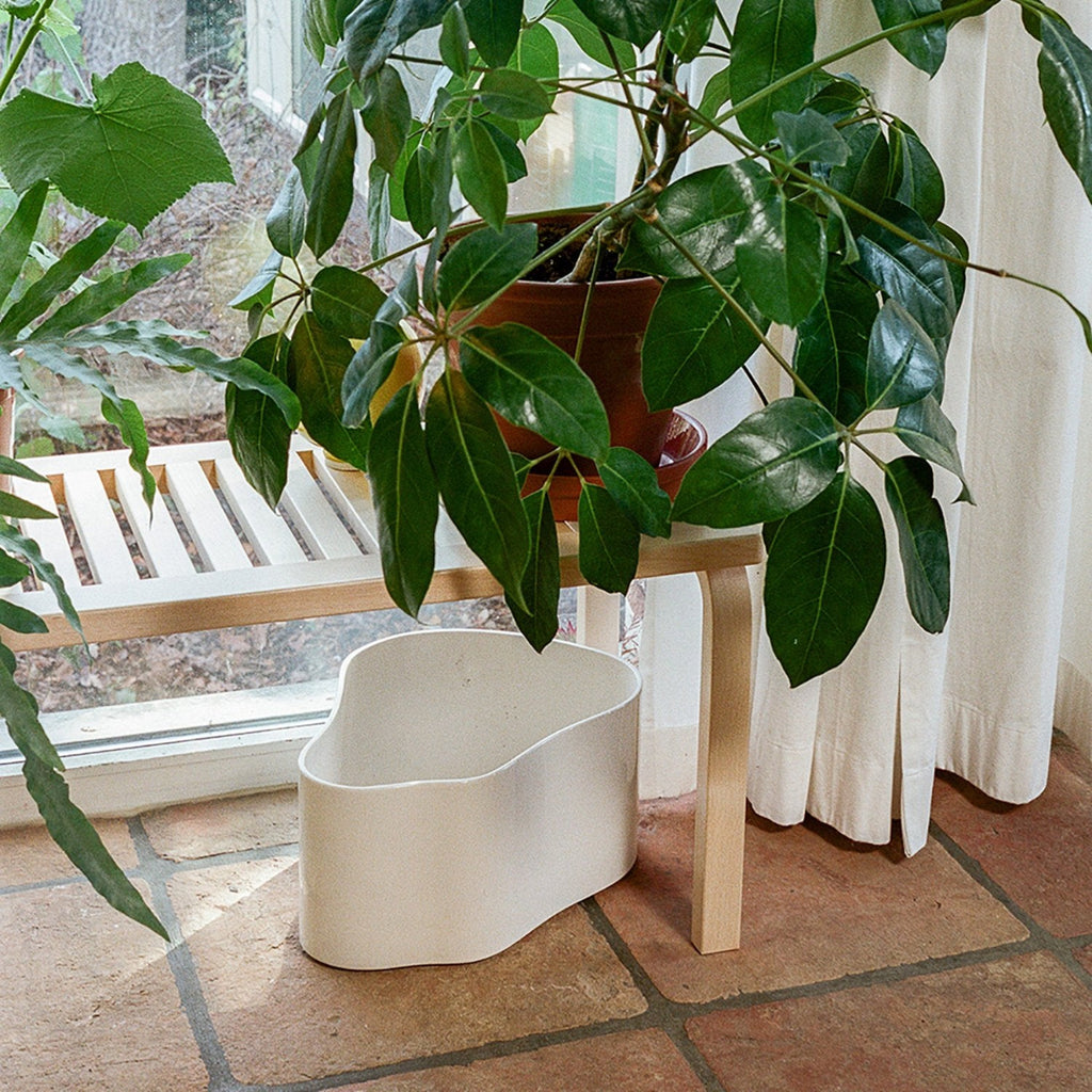 Interior view of a slatted Bench 153B with a clear lacquer finish. Plants are placed on top of the bench, while a large white Riihitie Plant Pot stands underneath.