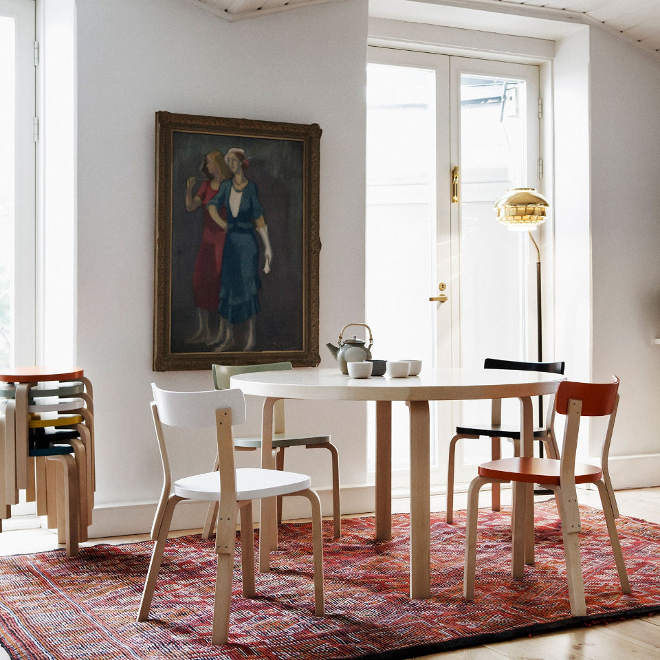 Dining room with round Artek Table 91 surrounded by four Chair 69, each chair featuring a differently painted seat and backrest. A red Moroccan rug lies beneath the dining group. In the room, a stack of Stool 60 with multicolored painted seats is visible. At the back of the space are a brass Floor Light A808.