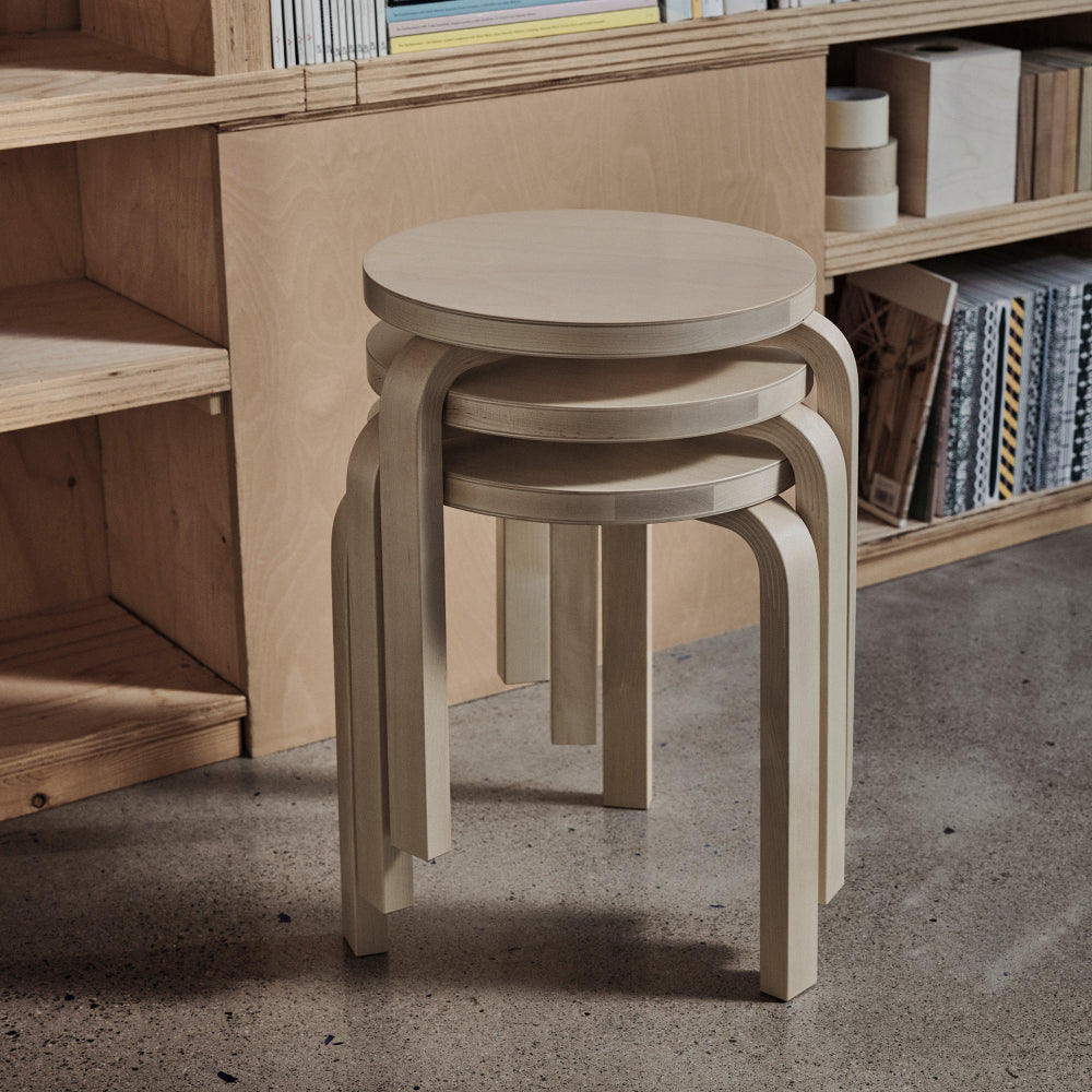 Three stacked Artek 60 Stools made of birch, with a bookshelf in the background.