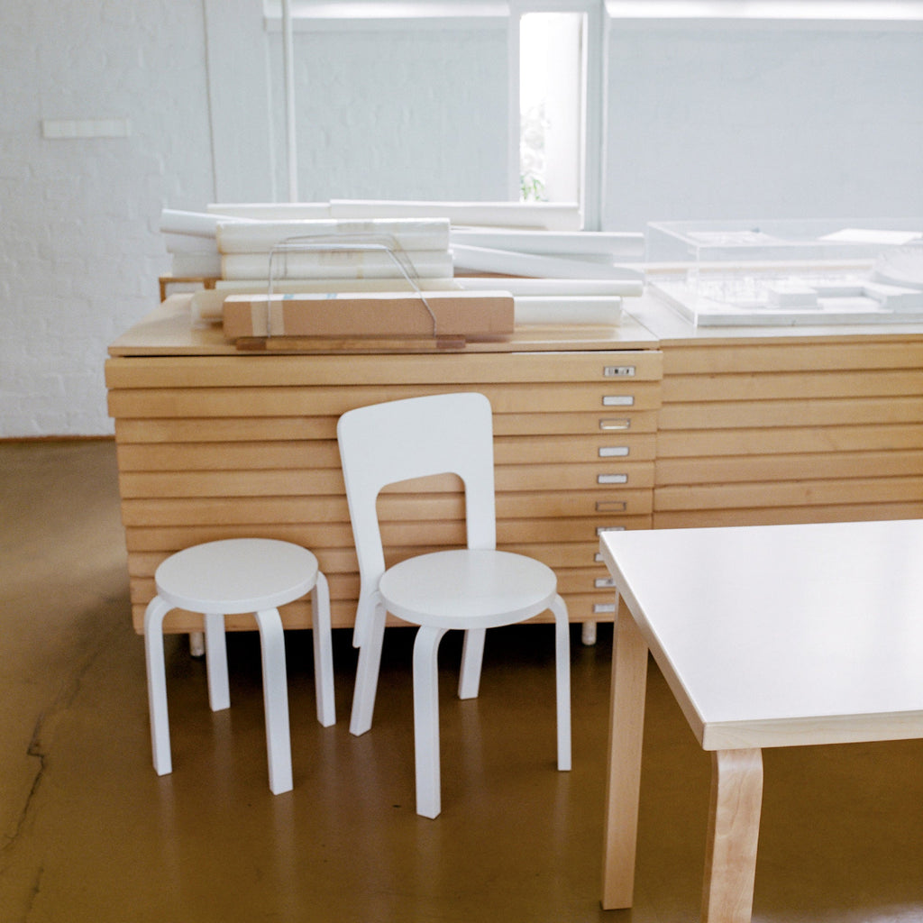 Interior view from Alvar Aalto Studio featuring a white brick wall with two white drawer units in front. A white lacquered Stool 60 and a white lacquered Chair 66 are placed in the foreground.