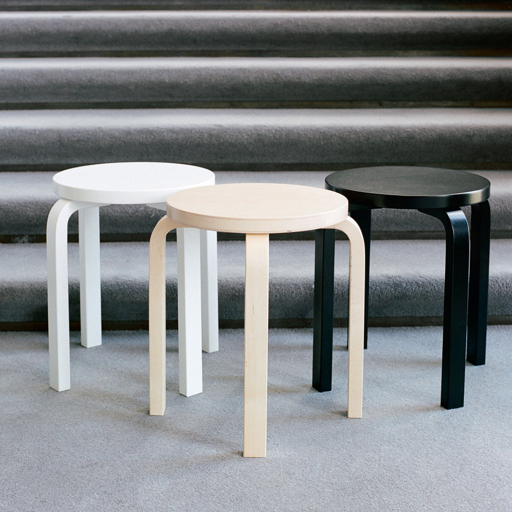 Three Artek E60 Stools placed side by side at the bottom of a staircase: painted white, lacquered birch, and painted black.