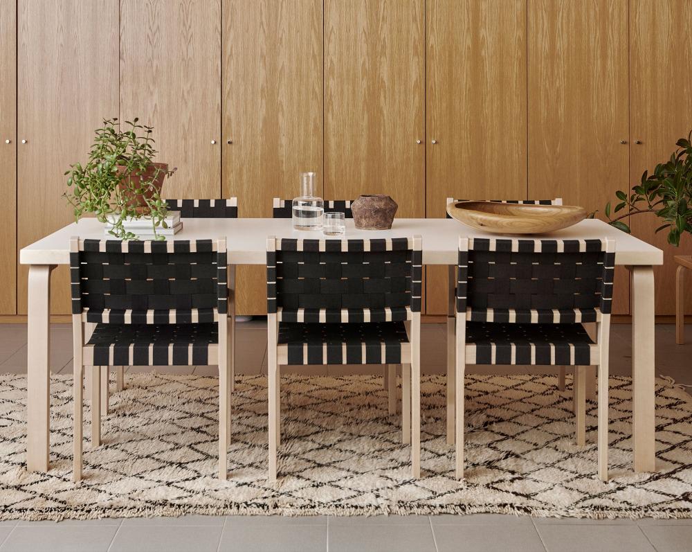 Dining area with Artek Table 86, surrounded by six Chair 611 placed along the long sides. Behind the table stands a wooden cabinet covering the entire wall. A Moroccan vintage rug lies beneath the table, and on the tabletop are a houseplant, a watering can, a glass, and two serving dishes.