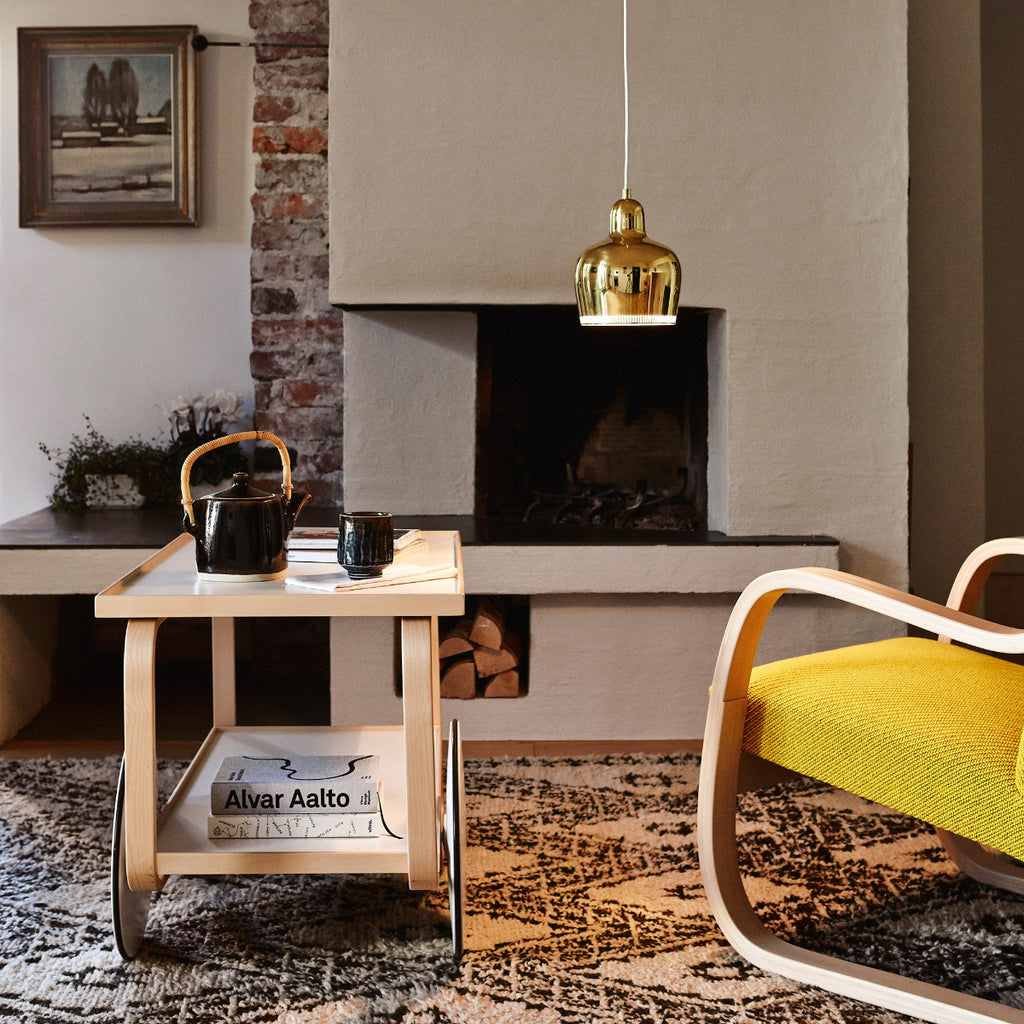 Interior view of a living room with a Tea Trolley 901 with a white laminate top placed in front of a fireplace. A yellow Armchair 402 stands nearby, while a brass A330S Pendant Light “Golden Bell” hangs in the centre of the room.