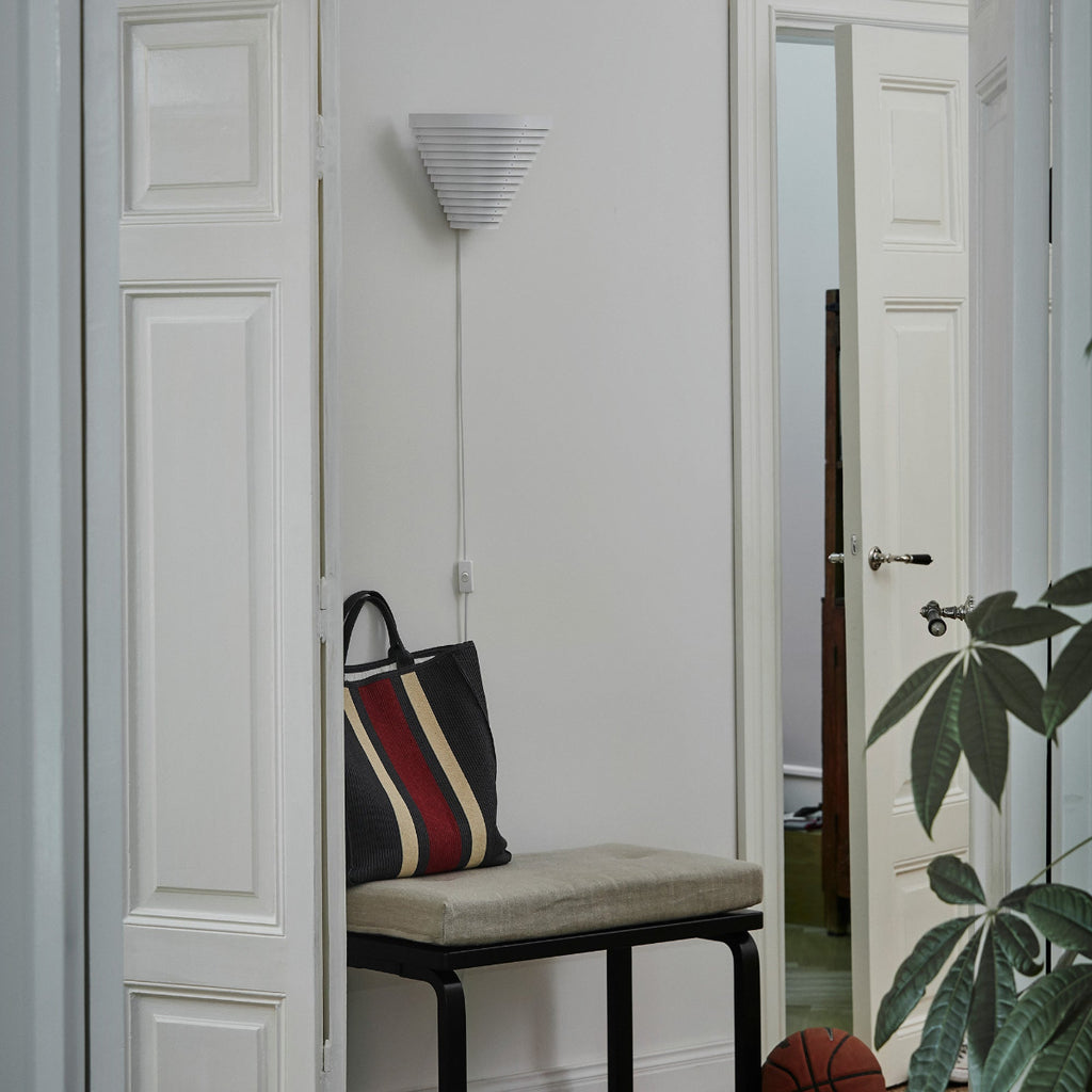 Interior view of a hallway lit by daylight featuring Wall Light A910 mounted on the wall above Bench 153B, positioned between two doors. The light is switched off.