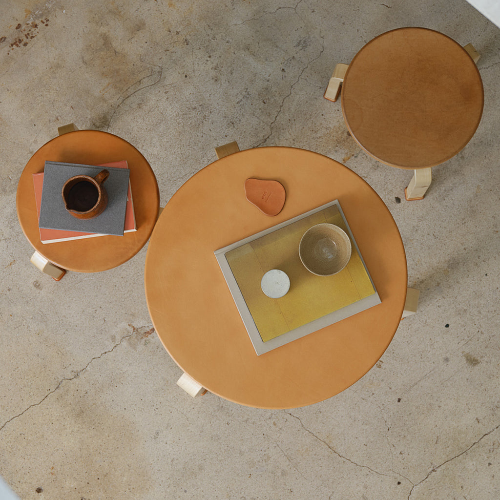 Top-down interior photograph of an Artek + Hender Scheme table with two stools. The table is positioned in the center of the image, with two stools placed around it. The leather-covered tabletop and stool seats have developed a beautiful brown patina over time. An Artek + Hender Scheme coaster, books, and cups are placed on the table, while a small stack of books rests on one of the stools.