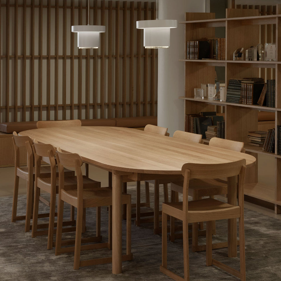 An oval table surrounded by eight oak Atelier Chairs. Two white Pendant Light A201 hang above the table. In this atmospheric dining space, a bookcase filled with old books and small objects stands beside the table.